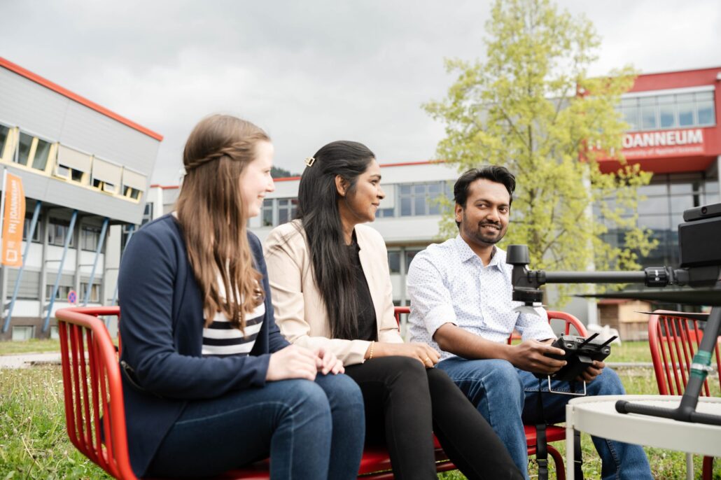 Partner descriptions image FHJ: Three students, 2 women, one man on a bench with a drone