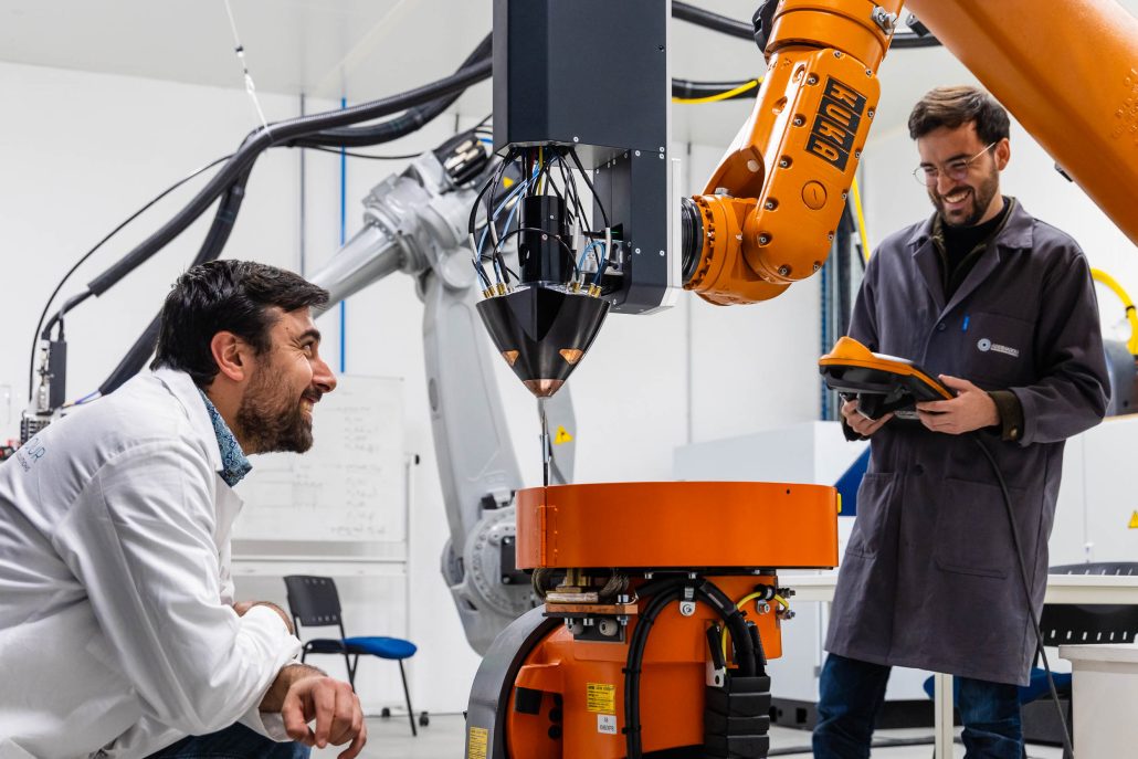 Two researchers in lab coats inspecting an industrial robot arm with laser deposition head in a university manufacturing lab